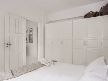 W sypialni ustawiono pojemne białe szafy wzdłuż jednej ściany. Śnieżna paleta barw wprowadza nieco chłodny nastrój, a...