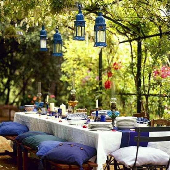 Niebieskie lataraenki, poduchy i dodatki w aranżacji biesiadnego stołu w letnim ogrodzie