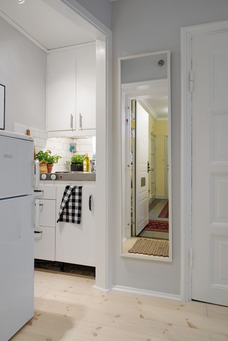 Białe szafki kuchenne ze stalowym blatem w mini aneksie kuchennym w otwartym widoku z przedpokojem z prostokatnym bialym lustrem