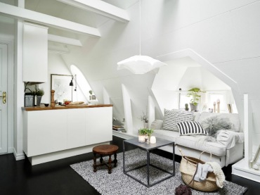 Otwarty salon z kuchnią , białe skośne ściany zbelkami,czarna podłoga,biała sofa na kółkach,metalowy stolik kawowy z drewnianym blatem,szary dywan i biało-czarne poduszki (27318)