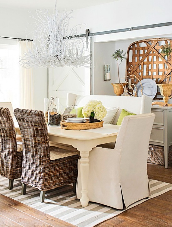Biały żyrandol z korzeni i witek nad drewnianym białym stołem w jadalni z wiklinowymi krzesłami,białe ubranka na krzesła,dywan w szaro-białe paski,szare szafki i drewniane dekoracje na ścianie