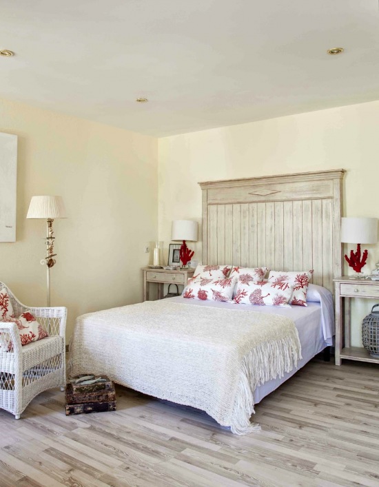 Drewniane bielone łóżko,biały wiklinowy fotel i parkiet w stylu vintage w sypialni z lampkami z czerwonego koralowca