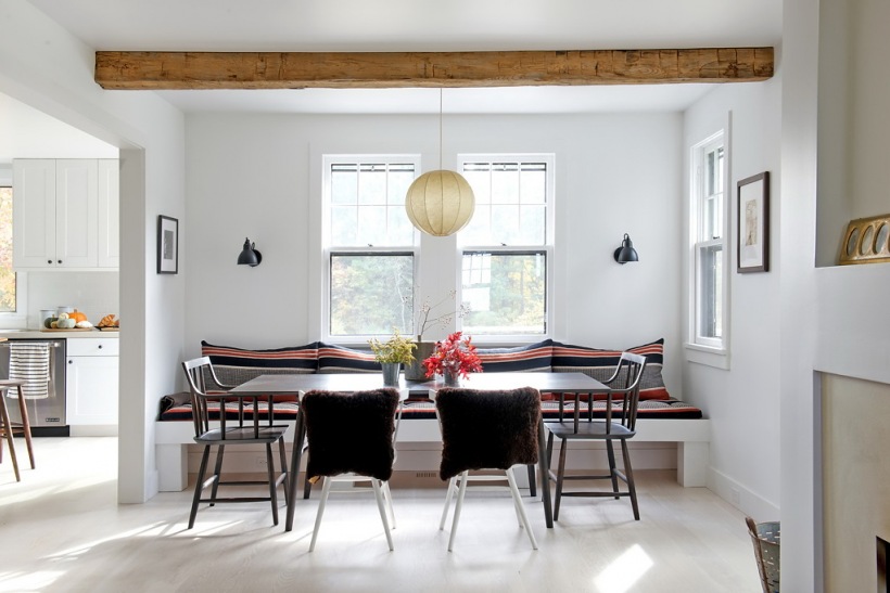 Biała jadalnia z drewnianymi belkami,murowanymi siedziskami z poduchami i różnymi krzesłami przy prostokątnym stole