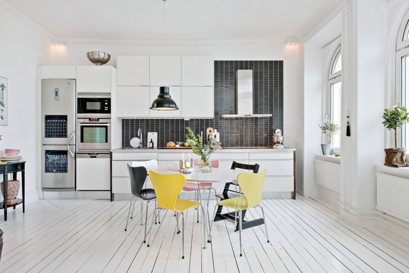 Biała kuchnia skandynawska z czarną glazurą na ścianie, okragłym stołem , żółtymi, czarnymi  i różowymi krzesłami