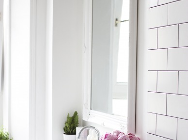 Dekoracyne, białe lustro nad umywalką nadaje aranżacji uroku, dodatkowo podkreślonego różowymi kwaitami w szklanym wazoniku. Białe ściany, sufit, szafka pod umywalką i płytki rozjaśniają wnętrze i sprawiają, że wydaje się higienicznie...