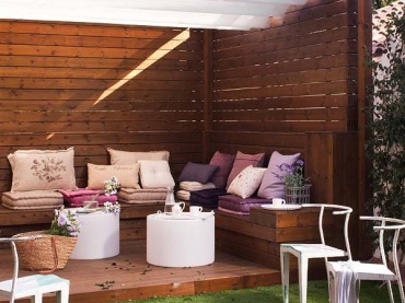 Drewniany brązowy taras z siedziskami, różowo-fioletowymi poduszkami i walcowatymi białymi stolikami (25518)