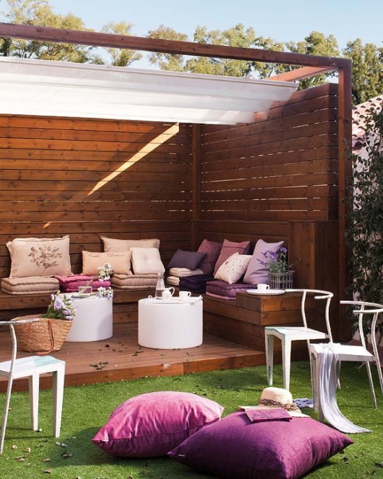 Drewniany brązowy taras z siedziskami, różowo-fioletowymi poduszkami i walcowatymi białymi stolikami