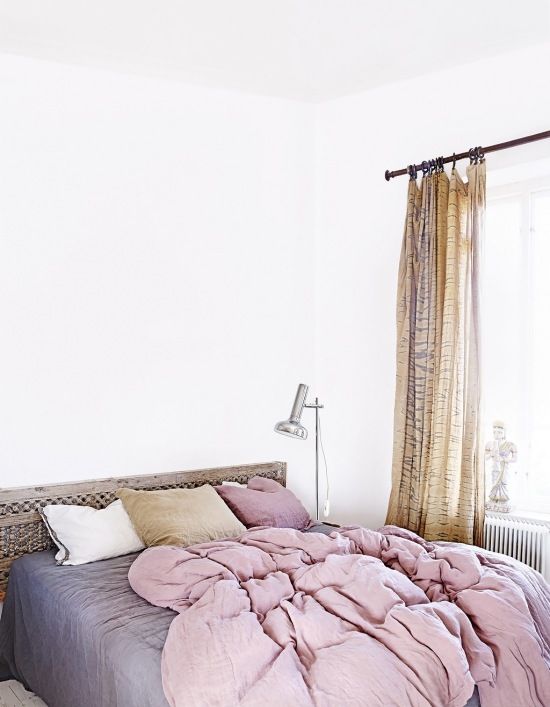 Lniane zasłony na drążkowym karniszu w białej sypialnu z marokańskim drewnianym łóżkiem i szaro-wrzosową pościelą