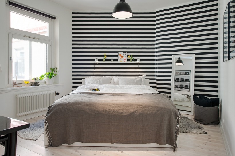 Tapeta w czarno-biale paski,stojące białe lustro i beżowa narzuta na łóżku w sypialni
