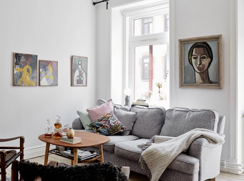 Kolorowe wspólczesne malarstwo w aranzacji białego salonu z szarą sofą i drewnianym okragłym stolikiem w stylu skandynawskim
