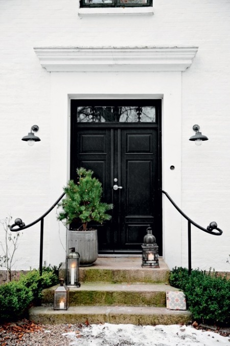 Białe ściany domu,czarne drzwi wejściowe,zielona choinka w szarym wazonie i metalowe latarenki na betonowych schodach