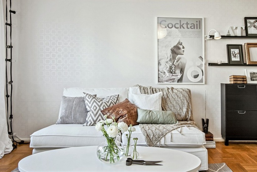 Stonowane pastelowe poduszki i pledy na białej sofie,czarna komoda i czarna girlanda z żarówek,srebrna tapeta na ścianie w salonie
