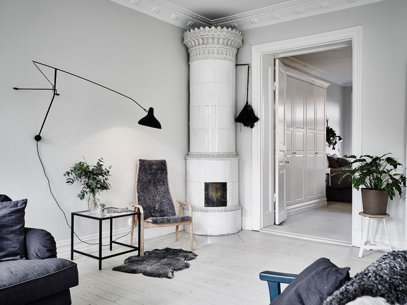 Biały ceramiczny piec w klasyczneym skandynawskim stylu,czarny kinkiet na wysięgniku,bielone deski z drewna na podłodze w salonie