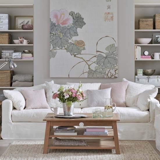 Drewniany tradycyjny stolik kawowy,szaro-różowy obraz z motywem roślin nym,jasnoszare ściany w wbudowanymi białymi pólkami i biała sofa na tkanym dywanie w salonie
