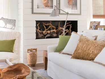 Białe panele z drewna na ścianie z kominkiem,dekoracje z korzeni,biala sofa z beżowymi i zielonymi poduszkami,wiklinowa skrzynia ze szklanym blatem w roli stolika w salonie (26161)