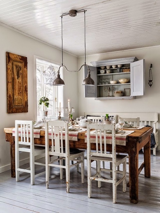 Miodowy drewniany stół z drewna w stylu rustykalnym,skandynawskie białe i patynowane krzesła vintage,szara wisząca podwójna lampa z metalu i szara szafka z pólkami na ścianie w kuchni wiejskiej w stylu skandynawskim
