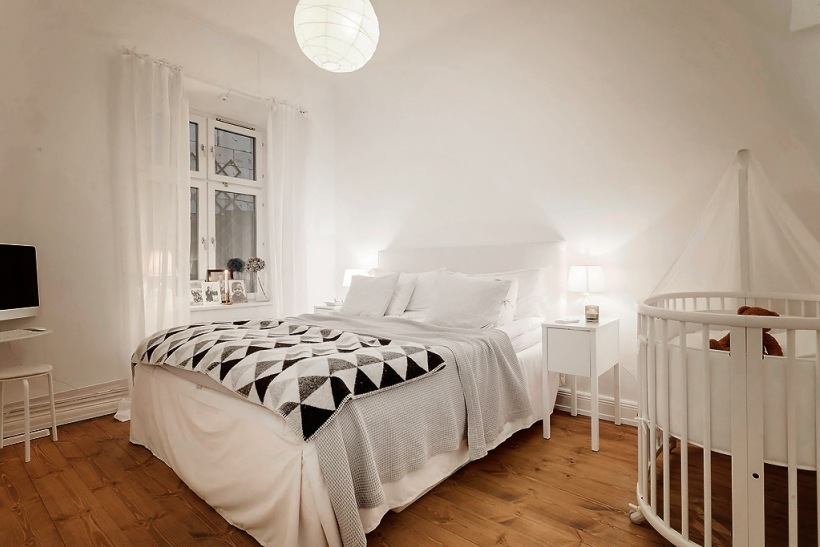 Biało-szaro-czarne trójkaty na skandynawskiej narzucie w białej sypialni małżeńskiej z drewnianym dziecięcym łóżeczkiem