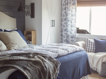 Niebieska narzuta do bezowego łóżka to idealna kombinacja w sypialni. Ściana z drewna w szarym kolorze , biała szafa i...