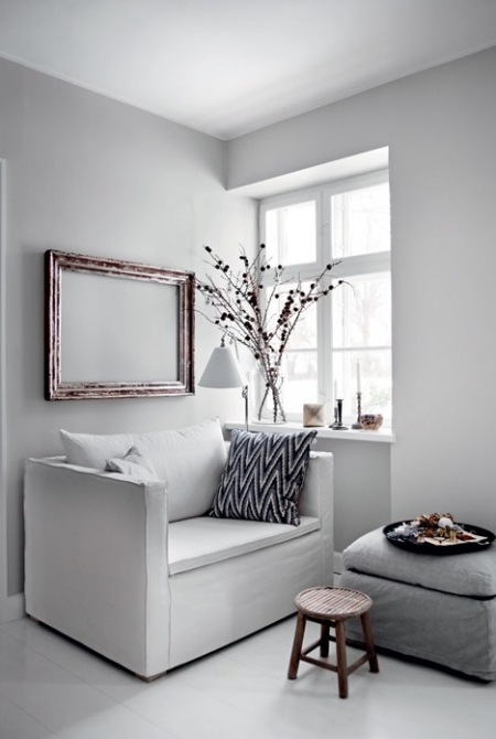 Minimalistyczna biała sofa z szarym podnóżkiem,drewniane ramy vintage i drewniany taboret w salonie przy oknie