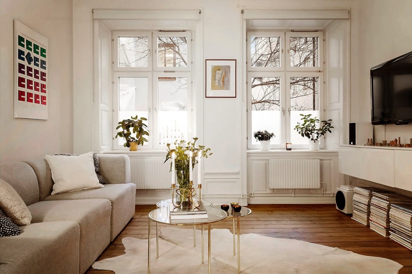 Nowoczesna sofa modułowa,okrągłe złote stoliki ze szklanymi blatami,wisząca biała szafka tv i biały dywan z bydlęcej skóry na drewnianej podłodze w salonie skandybnawskim