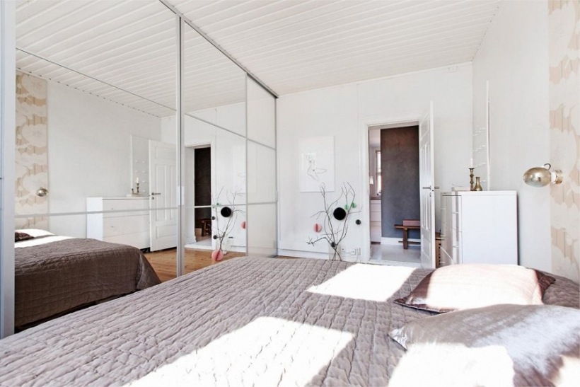 Aranżacja białej sypialni z szarą narzutą na łóżku i lustrzanymi szafami z przesuwnymi drzwiami