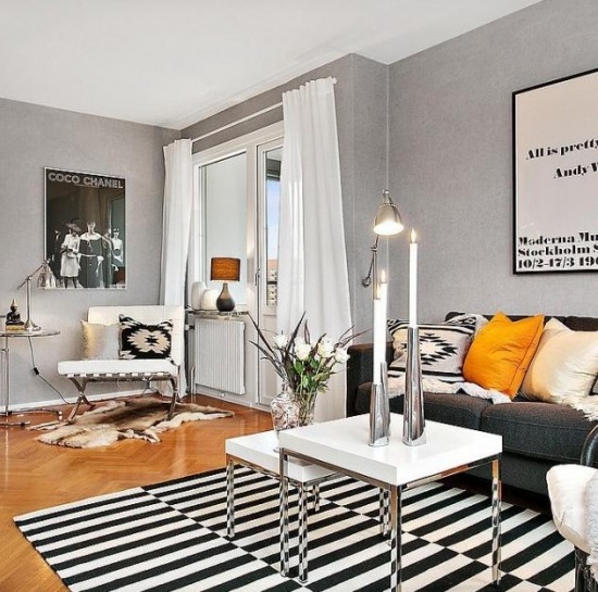 Szare ściany,białe stoliki nowoczesne ,biało-czarny dywan w paski i żółte poduszki na szarej sofie w salonie skandynawskim