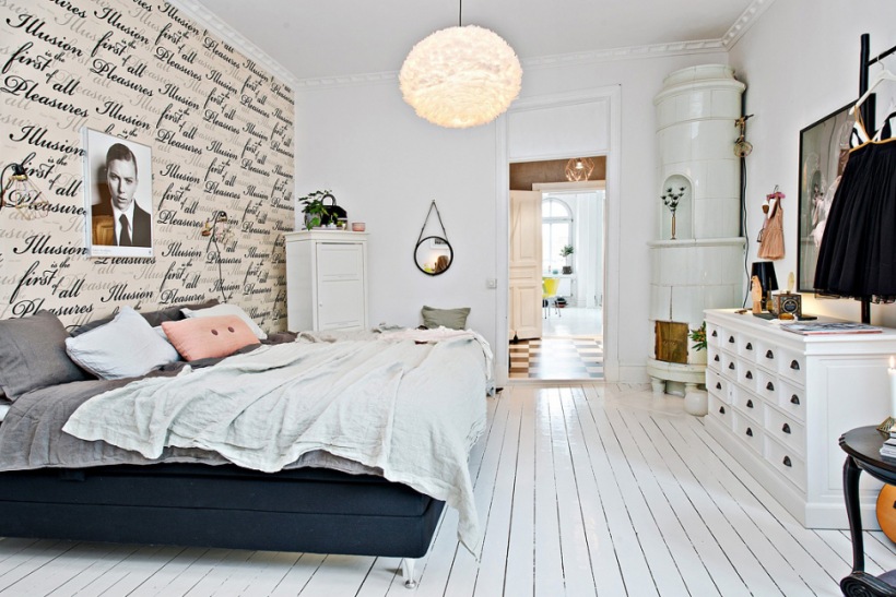 Drukowana tapeta z napisami,białe deski na podłodze,biała komoda szufladnik i czarne łóżko tapicerowane w skandynawskiej sypialni