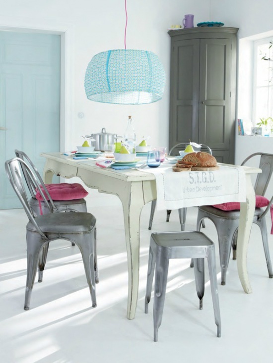 Prowansalski biały stół z drewna,szare krzesła i stołki industrialne z różowymi siedziskami,szara szafa i niebieska lampa nad stołem