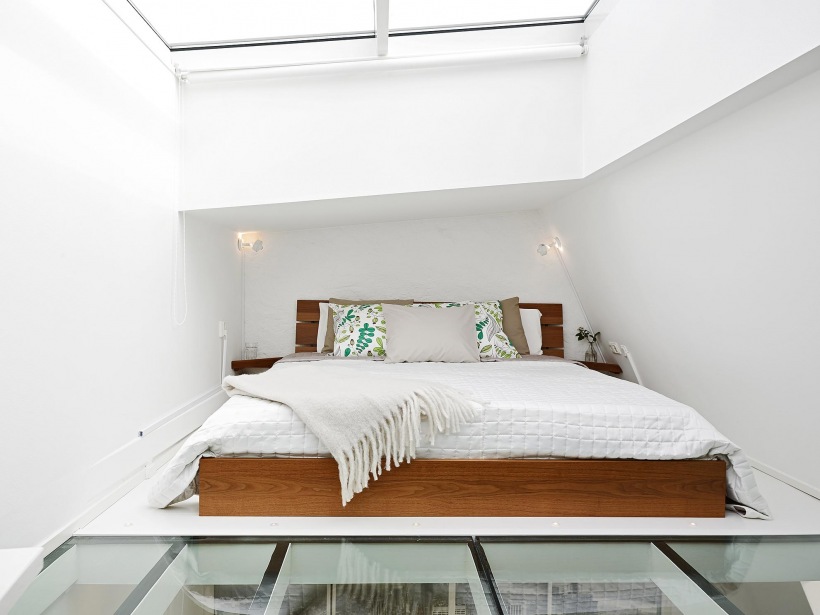 Drewniane łóżko w białej sypialni ze świetlikiem w suficie i przeszkloną podłogą