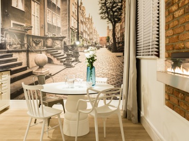 Fototapeta z ulicą Mariacką w Gdańsku stanowi tu tło dla jadalni i kuchni. Pomieszczenie utrzymane w kolorach brązowym,...