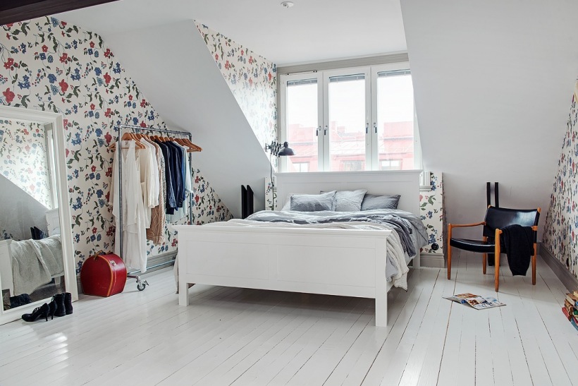 Białe łóżko,stojący metalowy wieszak na kółkach,drewniane ciemnobrązowe belki w sypialni,kwiatowa tapeta ścienna w niebiesko-czerwonym kolorze, duże białe lustro stojące