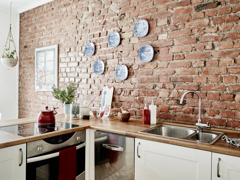 Ściana z czerwonej cegły,ozdobne biało-niebieskie talerze na ścianie  i biała kuchnia skandynawska z drewnianymi blatami