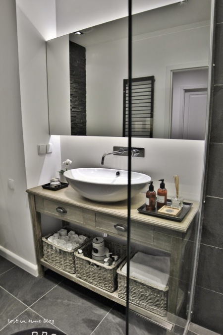 Owalna umywalka na konsolce z półką z bielonego drewna,duża tafla lustra nad umywalką,naścienna nowoczesna bateria,wiklinowe koszyki na ręczniki i akcesoria łazienkowe,szklana kabina z natryskiem