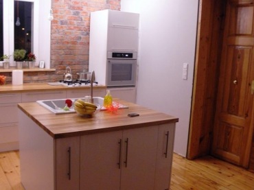 Białe szafki kuchenne w połączeniu z naturalnym drewnem i czerwonymi cegłami na ścianie. Jedyny akcent kolorystyczny...