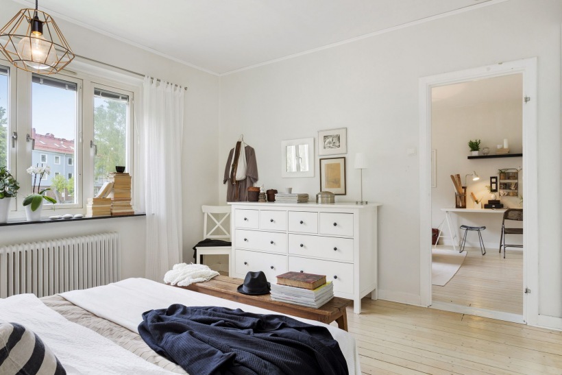 Biała  komoda z szufladami, druciana lampa i drewniana ławka z naturalnego drewna w dekoracji sypialni