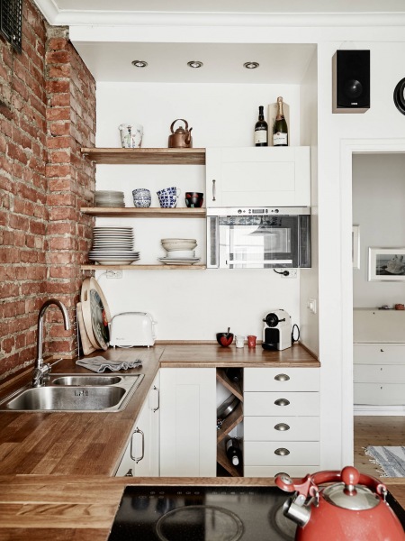 Biała kuchnia skandynawska z drewnianymi blatami i półkami i ścianą z czerwonej cegły