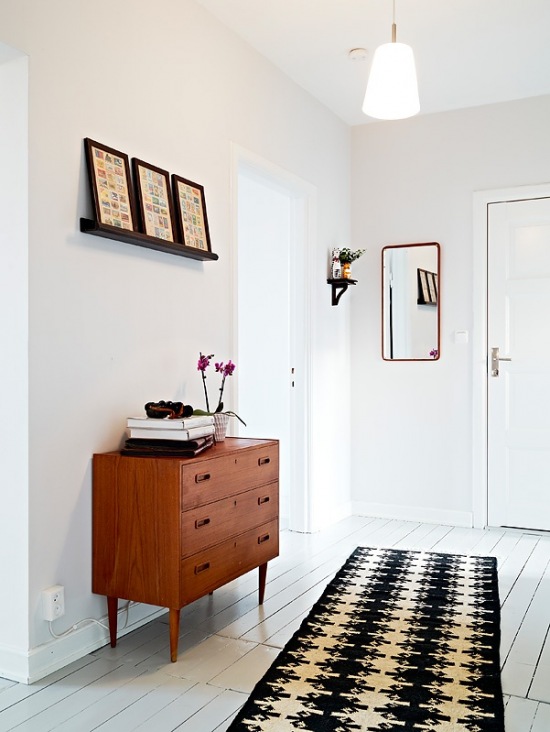 Podłoga z białych malowanych desek,czarna wąska półeczka z grafikami,drewniana komoda z szufladami i czarno-biały dywan w przedpokoju