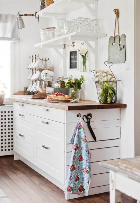 Skandynawska kuchenna komoda z drewnianym blatem,białe półki wiszące na ścianie, szklane i ceramiczne akcesoria w aranżacji kuchni