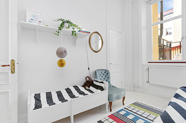 Biało-czarna pościel w paski,białe łóżko z drewna,kolorowy geometryczny dywanik i okrągłe lustro na ścianie w sypialni