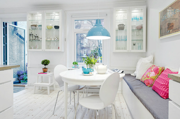 Wiszące białe witrynki, okrągły stół i ławka z poduszkami w skandynawskiej jadalni z niebieską lampą