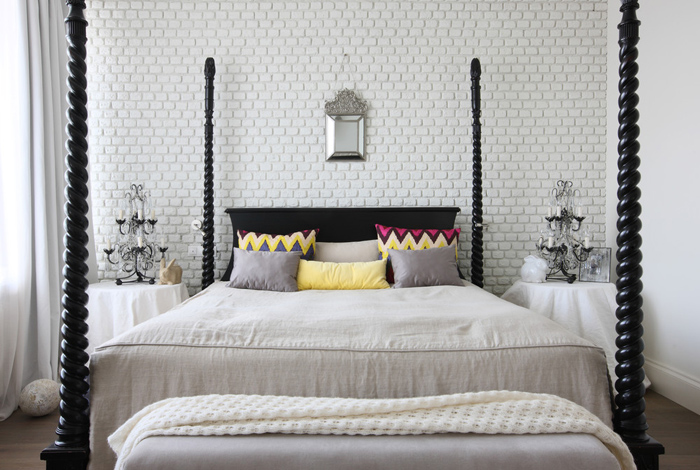Biała cegła na ścianie w sypialni ze stylowym drewnianym łóżkiem,kolorowymi poduszkami w zygzaki i stylowymi świecznikami lampami na stolikach