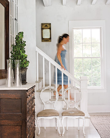 Stylowy szufladnik z marmurowym  blatem i gustawiańskie krzesła w domowym bialym korytarzu ze schodami