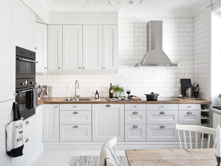 Biała kuchnia skandynawska ze srebrnymi uchwytami w kształcie muszli,drewnianymi blatami i stalowym okapem i płytką glazurowaną białą cegiełką na ścianie