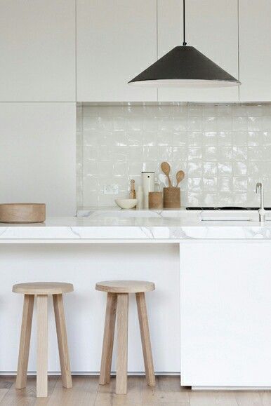 Białe nowoczesne szafki kuchenne,drewniane taborety,biała glazurowana płytka na ścianie i marmurowe blaty w minimalistycznej kuchni