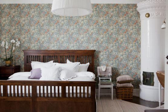 Kolonialne drewniane łóżko,ceramiczny skandynawski piec i tapeta z wmotywem kwiatowym w aranżacji sypialni