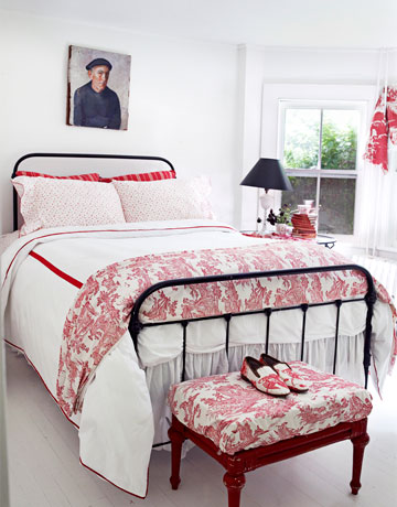 Czarne kute łóżko,czerwony puf i stylowa biało-czerwona pościel w wiejskiej sypialni