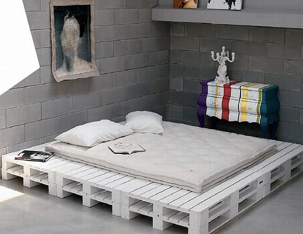 Podstawa i ramy łóżka z białych drewnianych palet w awangardowej sypialni z betonowymi scianami i kolorową komodą