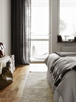 Ciemnoszare lniane zasłony,czarna narzuta z białymi szwedzkimi wzorami,biały futrzak na podłodze w dekoracji sypialni