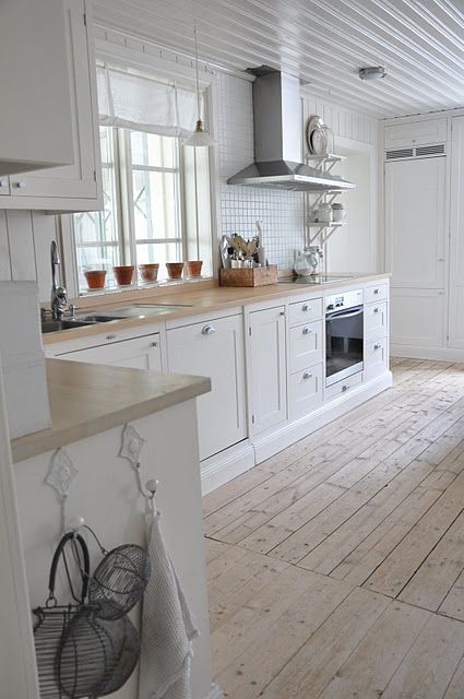 Podłoga z drewnianych desek w naturalnym kolorze, białe szafki kuchenne z drewnianymi blatami i biała drobna płytka glazurowana na ścianie