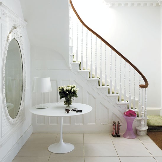 Owalne białe lustro,okragły stolik na jednej nodze i toczone białe tralki przy drewnianych schodach